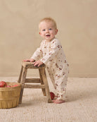 A baby standing on a woven rug next to a small wooden stool, wearing a cream-colored long-sleeve footless onesie with a brown floral pattern. A wooden basket of red apples sits on the floor to the left.