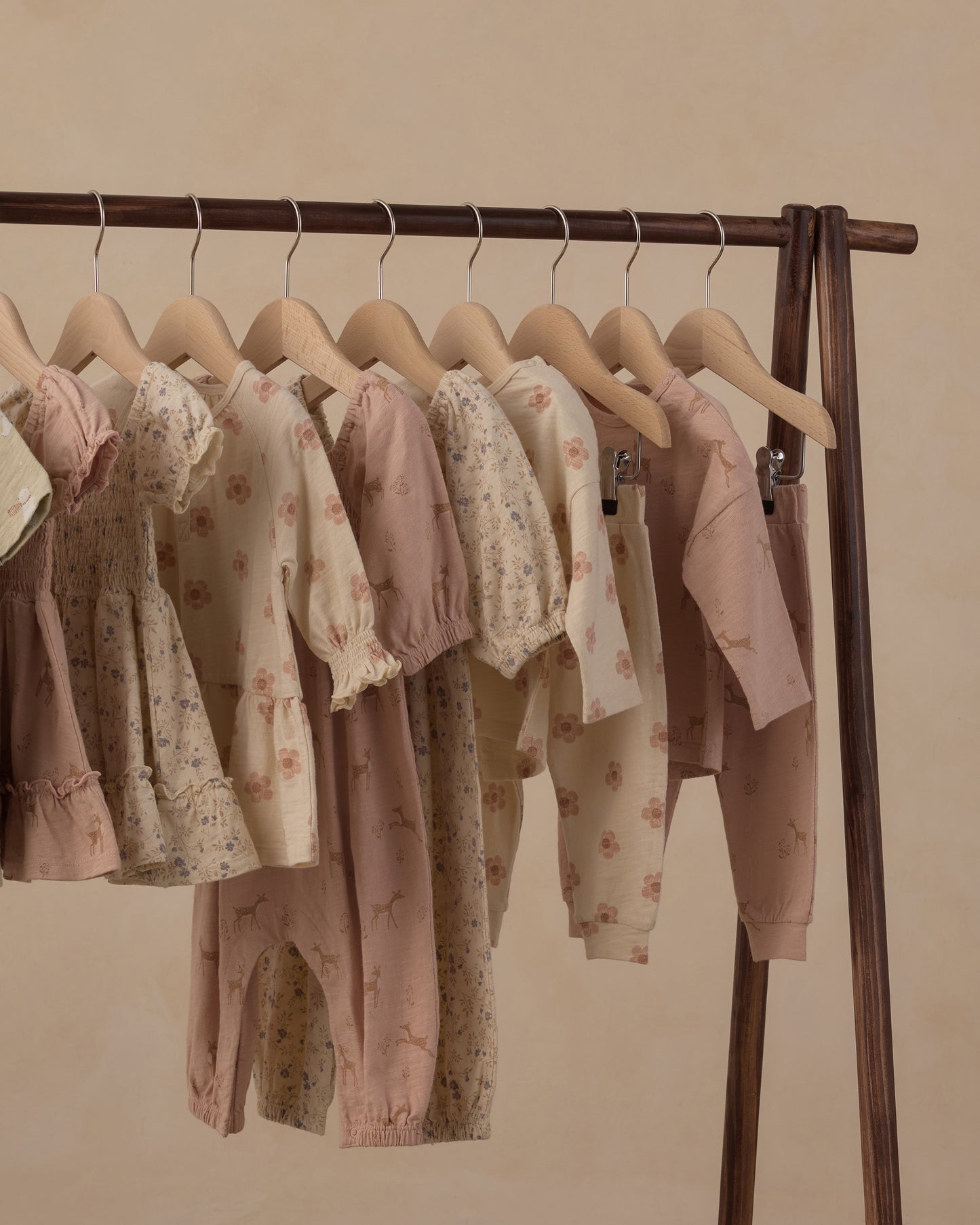 A set of baby clothing in soft pastel colors, including floral long-sleeve tops and matching pants, hanging on wooden hangers from a dark wood clothing rack against a warm beige background.