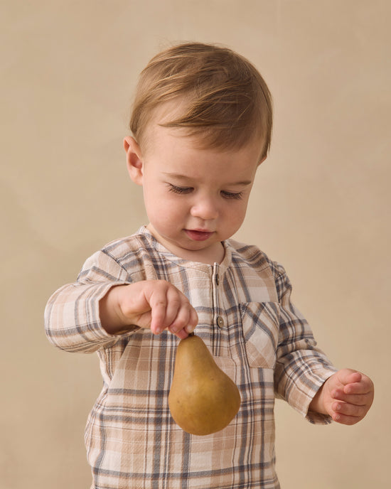 A toddler boy wearing a beige and brown plaid long-sleeve shirt holds a brown pear against a warm beige studio background.