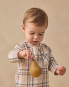 A toddler boy wearing a beige and brown plaid long-sleeve shirt holds a brown pear against a warm beige studio background.