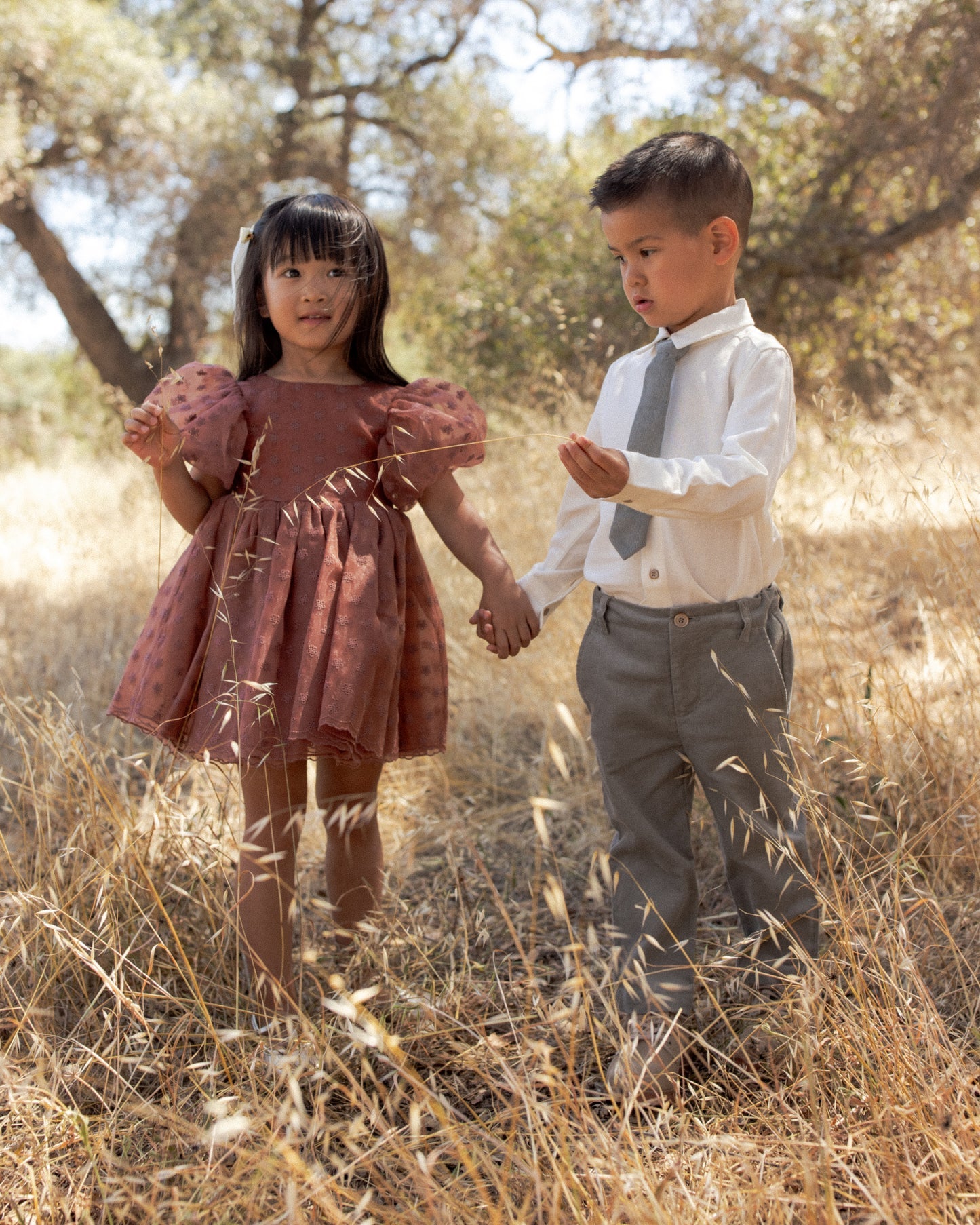 A young girl in a dusty rose puff-sleeve dress and a boy in a white shirt with a gray tie and gray trousers, holding hands and walking through dry grass outdoors.