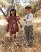 A young girl in a dusty rose puff-sleeve dress and a boy in a white shirt with a gray tie and gray trousers, holding hands and walking through dry grass outdoors.