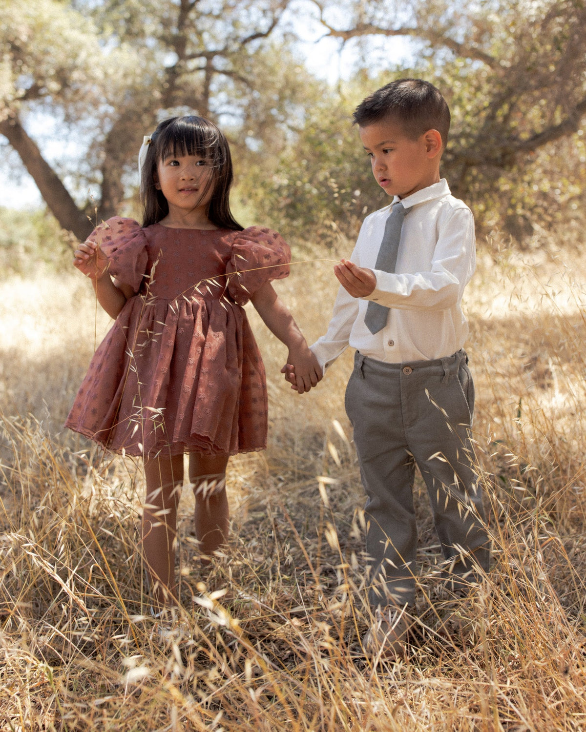 A young girl in a dusty rose puff-sleeve dress and a boy in a white shirt with a gray tie and gray trousers, holding hands and walking through dry grass outdoors.
