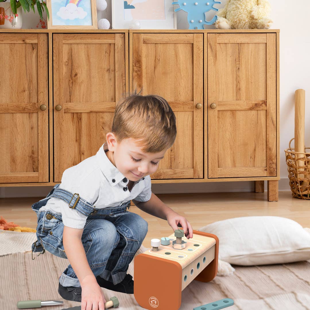 A young boy in a white shirt and denim overalls kneels on a light rug, playing with a wooden toy block set on an orange base in a bright room with wooden cabinets.