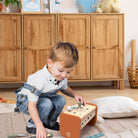 A young boy in a white shirt and denim overalls kneels on a light rug, playing with a wooden toy block set on an orange base in a bright room with wooden cabinets.