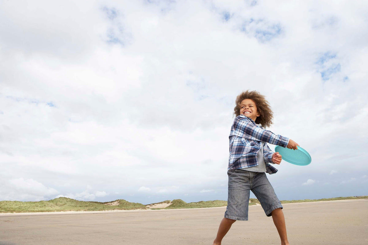A child smiling and throwing a bright turquoise flying disc across a sandy beach under a cloudy sky.