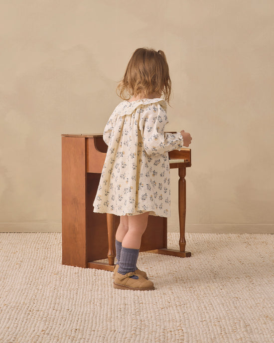 A toddler stands with their back to the camera, wearing a cream-colored long-sleeve dress with a delicate blue floral print, gray socks, and tan shoes, standing next to a small wooden table in a warm, neutral-toned room.