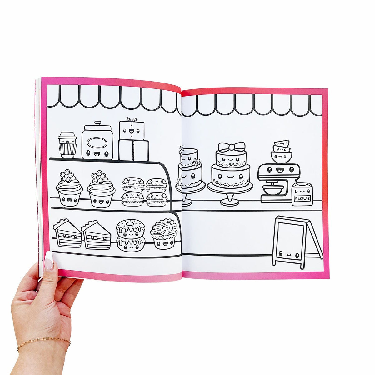 Coloring book page with cute kitchen and cake illustrations held open by a hand.