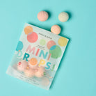 Packaging of 'Mini Drops' with colorful bath bombs on a light blue background