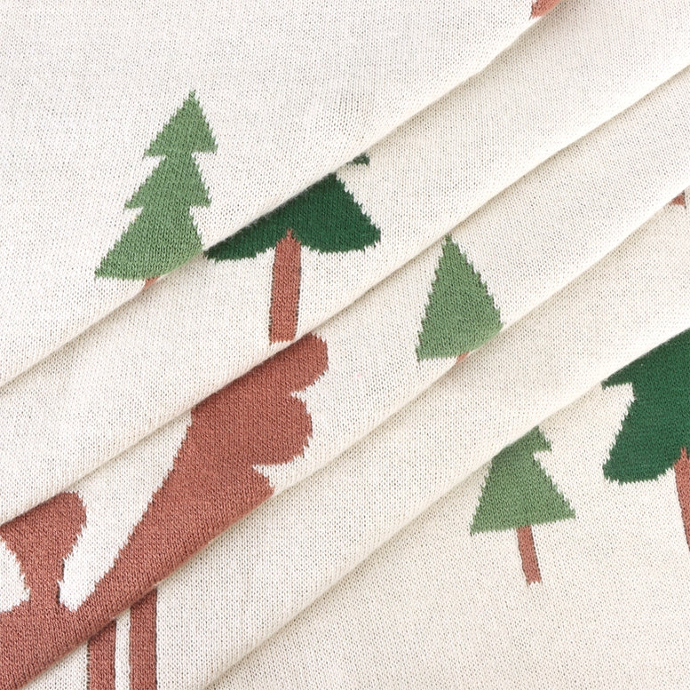 A close-up of a cream-colored fabric with a repeating pattern of green pine trees and reddish-brown deer silhouettes, arranged in soft diagonal folds.
