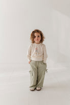 Child wearing a beige sweater and green pants standing against a white background