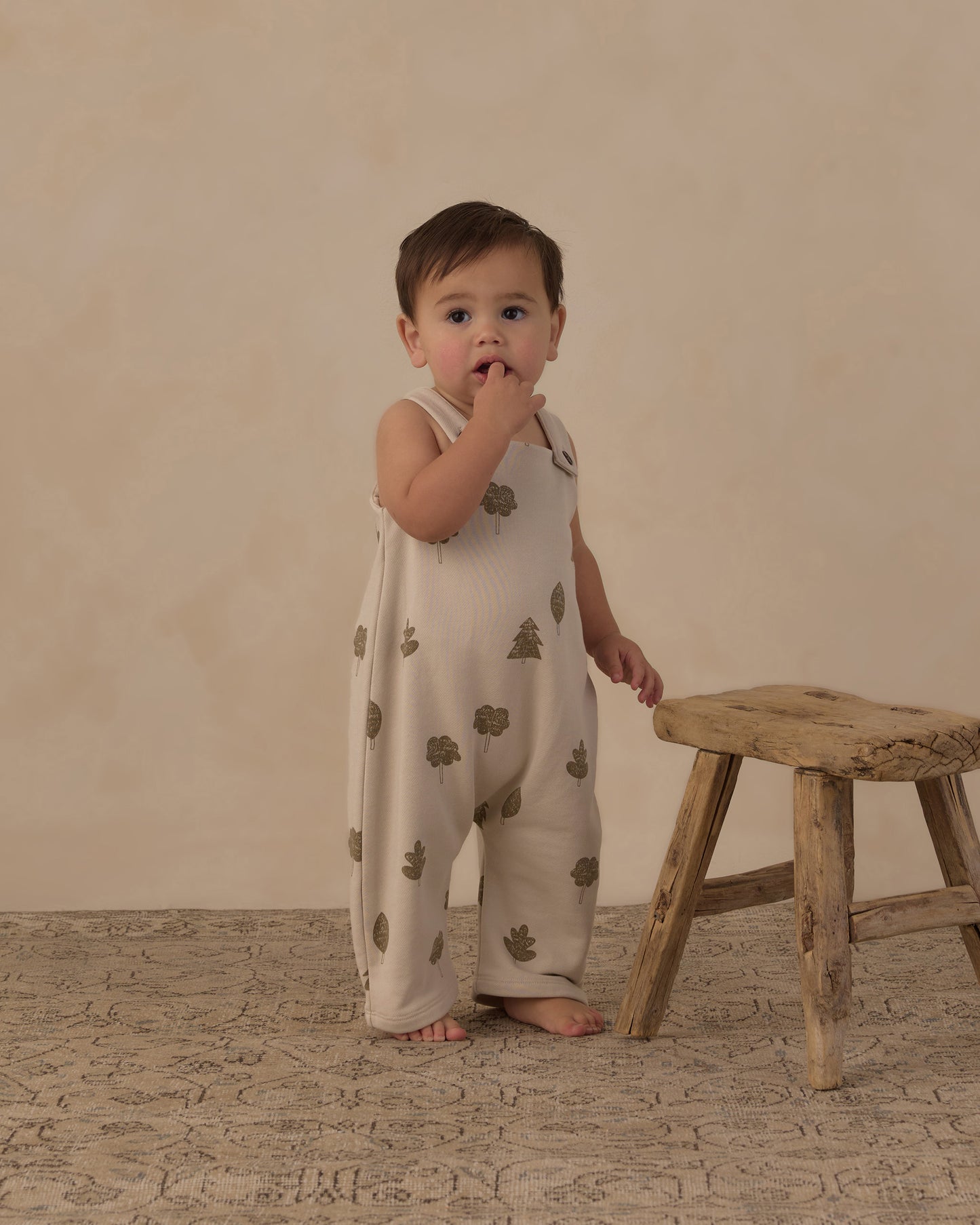 A baby standing on a textured rug next to a rustic wooden stool, wearing a cream-colored sleeveless romper with a green tree pattern, set against a warm beige studio backdrop.