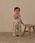 A baby standing on a textured rug next to a rustic wooden stool, wearing a cream-colored sleeveless romper with a green tree pattern, set against a warm beige studio backdrop.