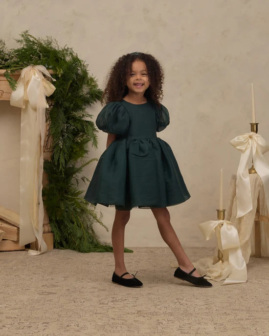 A young girl with curly dark hair stands in a studio setting wearing a dark teal puff-sleeve dress with black flats. The background features a textured beige wall, a green garland with a cream ribbon on the left, and a decorative stand with a cream ribbon and candle on the right.
