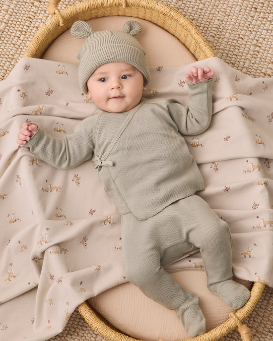 A baby wearing a sage green knit footed one-piece outfit with a matching beanie hat lies on a beige blanket printed with small deer, inside a round woven basket.