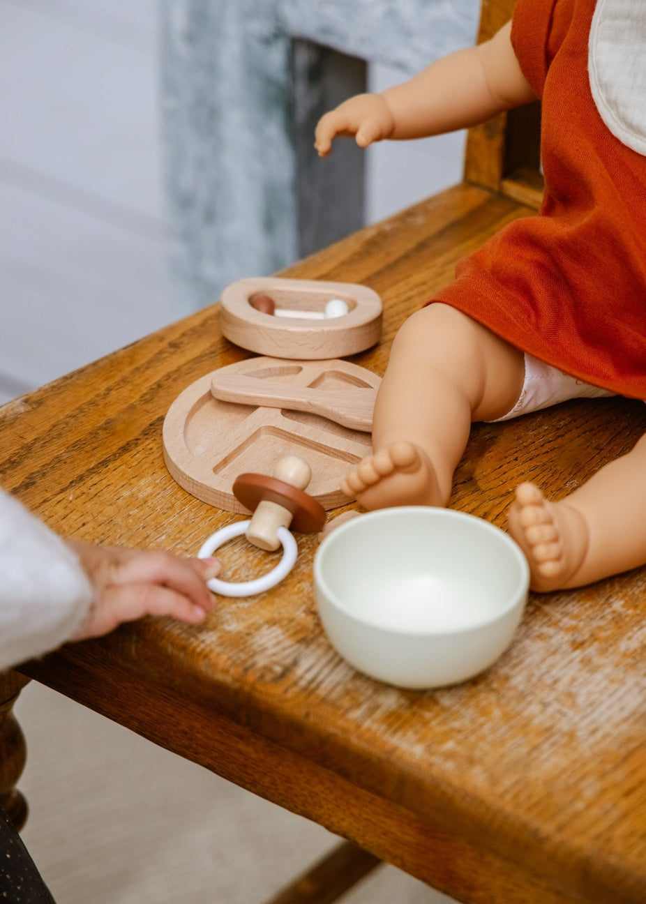 A close-up of a wooden baby sensory toy set on a rustic wooden table, featuring a wooden rattle, a wooden teething ring with a white ring, a small wooden spoon, and a white ceramic bowl. A doll in an orange dress and a baby's hand are interacting with the toys.