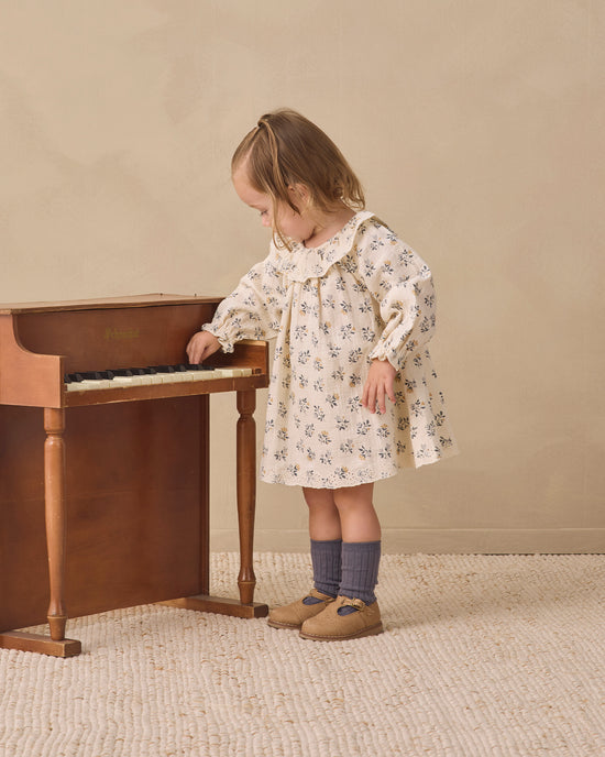 A young child wearing a cream-colored long-sleeve dress with a delicate blue floral pattern, standing next to a wooden piano in a warm, neutral-toned room.