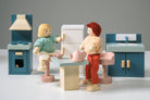 Wooden kitchen furniture set by Mentari against a white background, featuring two wooden dolls seated at a light wood-colored table with a teapot and two cups, surrounded by a blue stove with an oven and hood, a white refrigerator with wooden handles, and a blue sink unit with a cupboard.