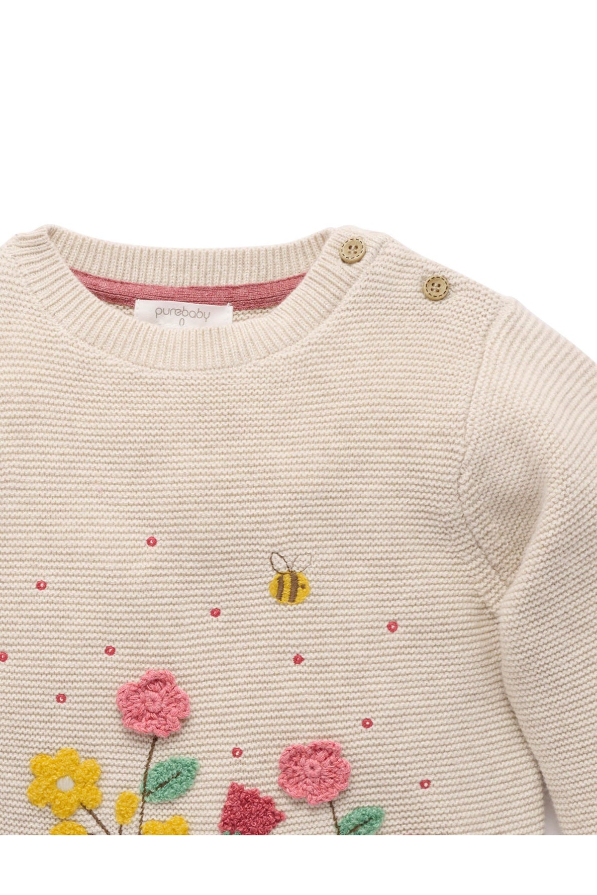 Close-up of a cream knit baby sweater with embroidered pink and yellow flowers, green leaves, a small bee, and pink dots, plus gold-toned shoulder buttons.
