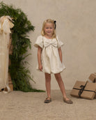 A young blonde girl stands in a studio setting wearing a short cream dress with a large bow at the chest, flutter sleeves, and leopard-print flats. A beige textured backdrop, greenery, and wrapped gifts are visible in the background.