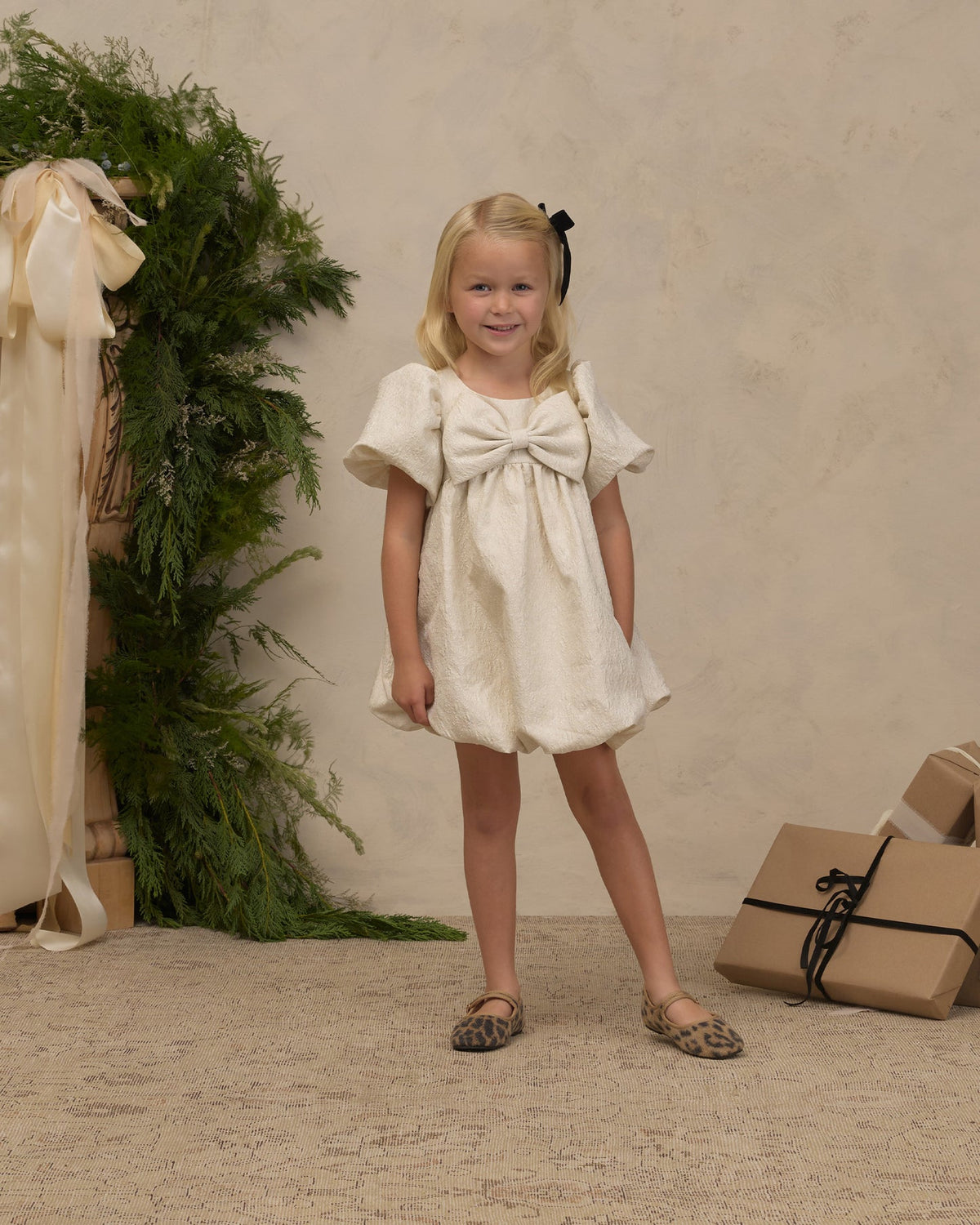 A young blonde girl stands in a studio setting wearing a short cream dress with a large bow at the chest, flutter sleeves, and leopard-print flats. A beige textured backdrop, greenery, and wrapped gifts are visible in the background.