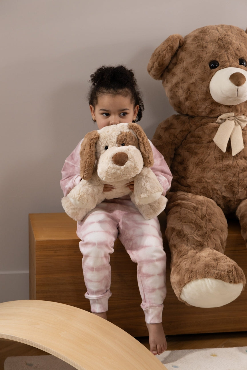Child in pajamas holding a stuffed dog next to a large teddy bear on a wooden surface.