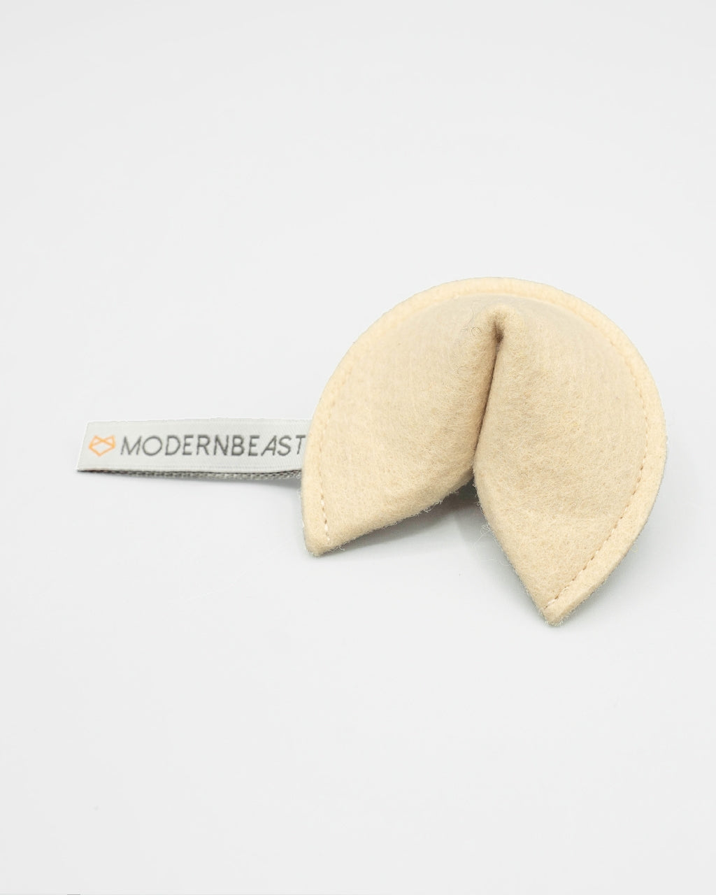 A cream-colored felt baby teething toy shaped like a fortune cookie, with a white fabric tag on the left side printed with the brand name 'MODERNBEAST'.