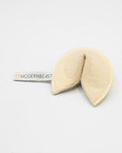 A cream-colored felt baby teething toy shaped like a fortune cookie, with a white fabric tag on the left side printed with the brand name 'MODERNBEAST'.