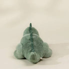 A plush dinosaur toy in a dusty sage green color, photographed from behind against a neutral, light beige background.