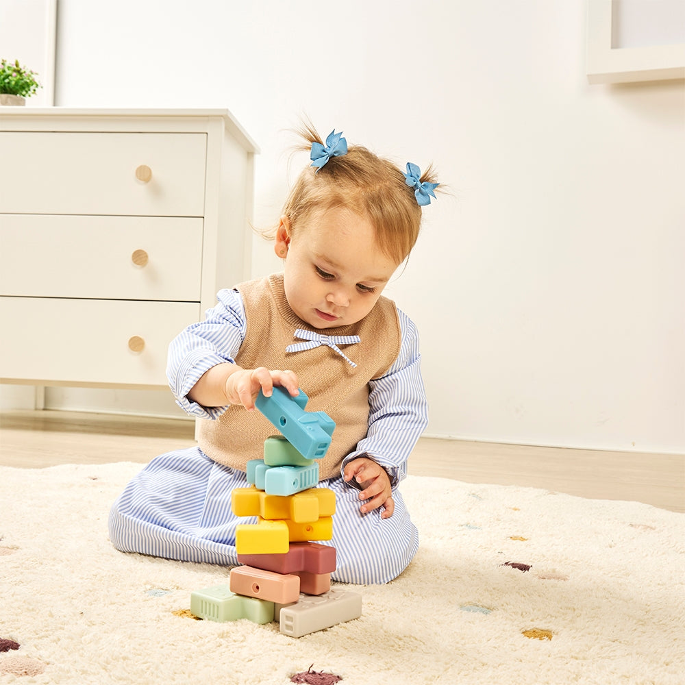 A toddler girl with blue hair bows sits on a cream rug, stacking pastel wooden blocks. She wears a blue and white striped long-sleeve top with a beige vest.