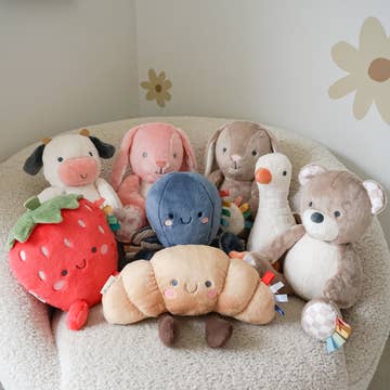 Collection of soft toys including a strawberry, dog, rabbit, blue creature, caterpillar, goose, and bear on a cushion.