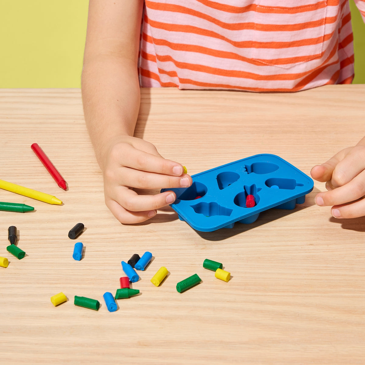 Child playing with colorful crayon pieces and a blue mold on a wooden table.