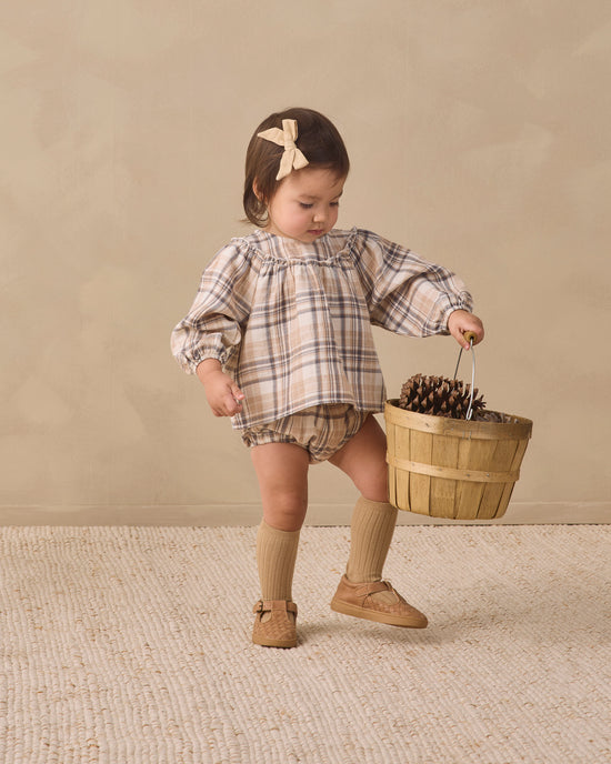 A toddler girl wearing a beige and brown plaid long-sleeve romper with matching knee-high socks and tan shoes, holding a small wooden basket on a textured cream rug against a warm beige studio backdrop.