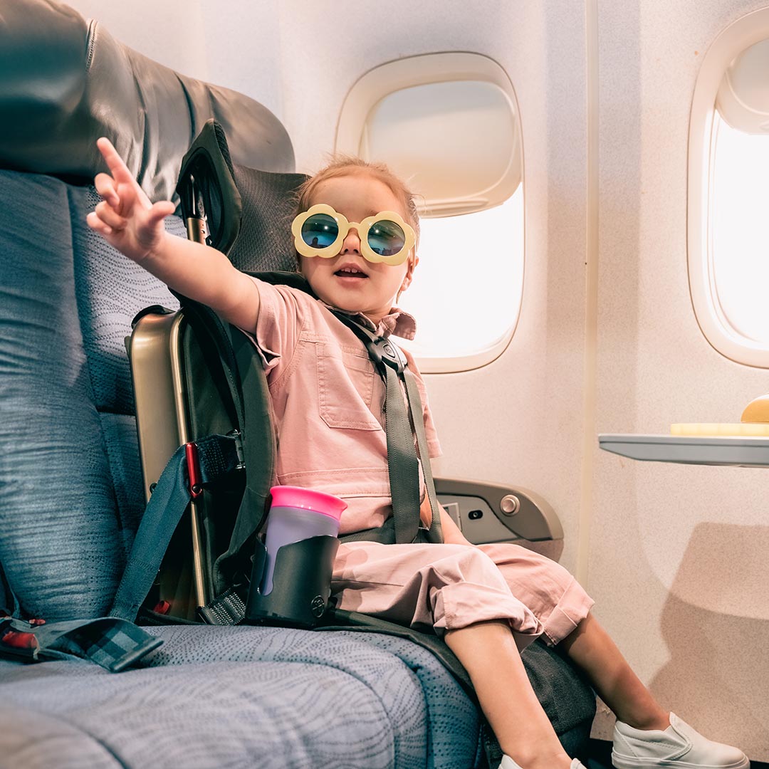 Child wearing sunglasses sitting in an airplane seat with a suitcase next to them.