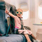 Child wearing sunglasses sitting in an airplane seat with a suitcase next to them.