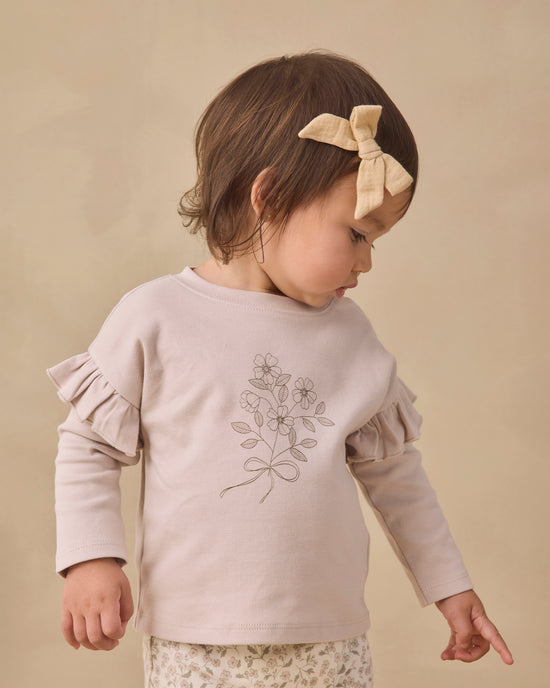 A toddler girl wearing a pale blush pink long-sleeve top with ruffled sleeves and a delicate floral line-art print on the front, styled with a beige bow headband against a warm beige background.