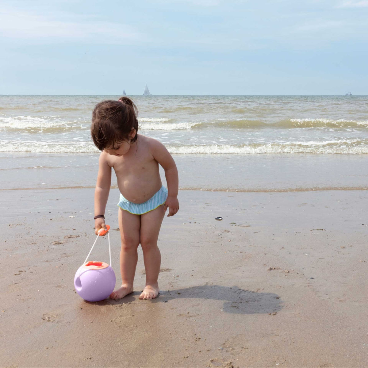 Child playing on a beach with a toy ball, wearing a light blue diaper.