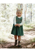 A young child stands on a forest path wearing a dark green pinafore-style dress over a white long-sleeve top, with embroidered floral details on the chest, brown boots, and hair styled in two buns.