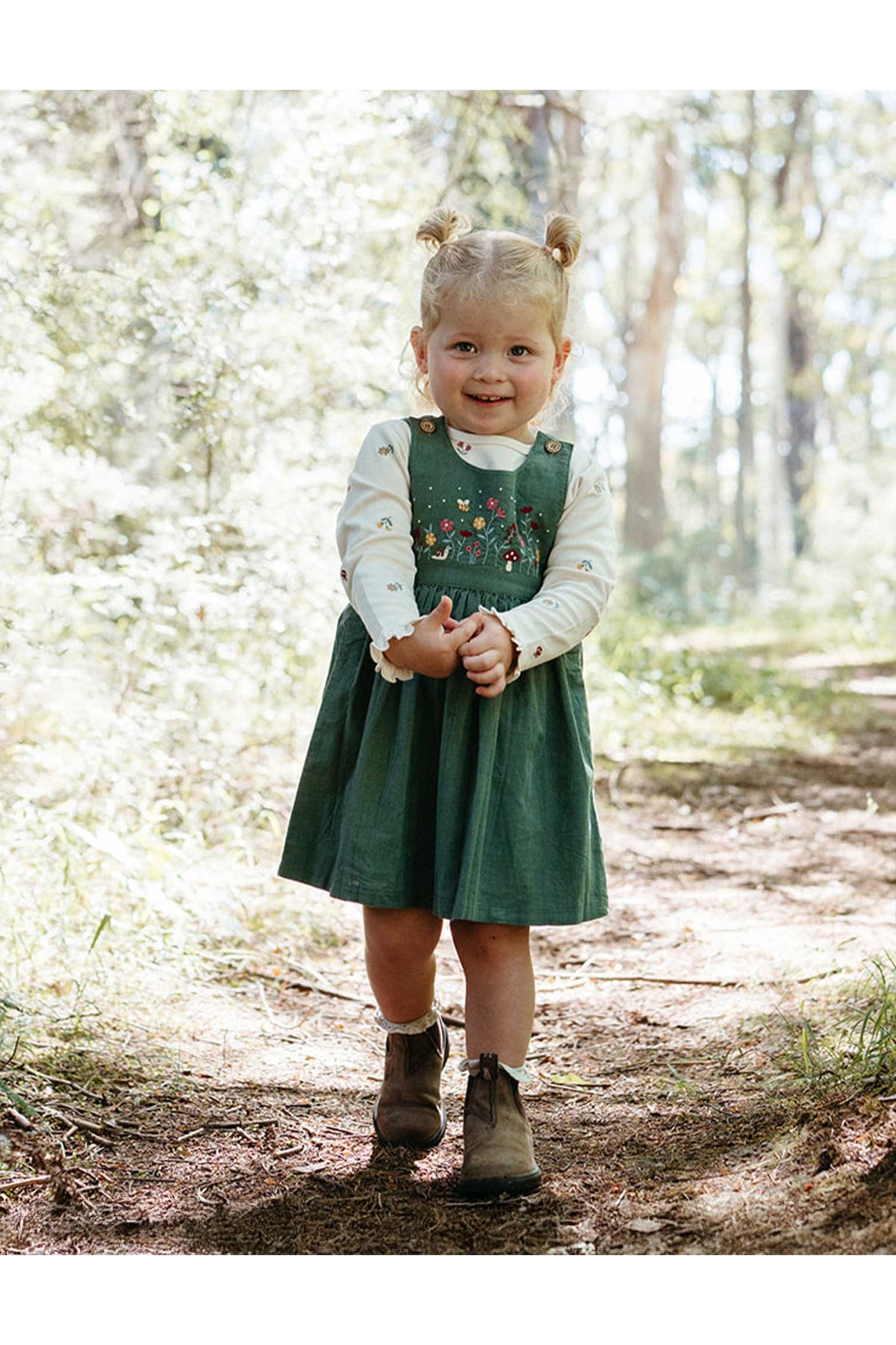 A young child stands on a forest path wearing a dark green pinafore-style dress over a white long-sleeve top, with embroidered floral details on the chest, brown boots, and hair styled in two buns.