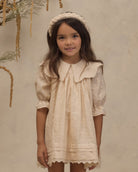 A young girl with long brown hair wearing a cream-colored lace dress with a large Peter Pan collar, puff sleeves, and a matching braided headband, standing against a textured beige wall.