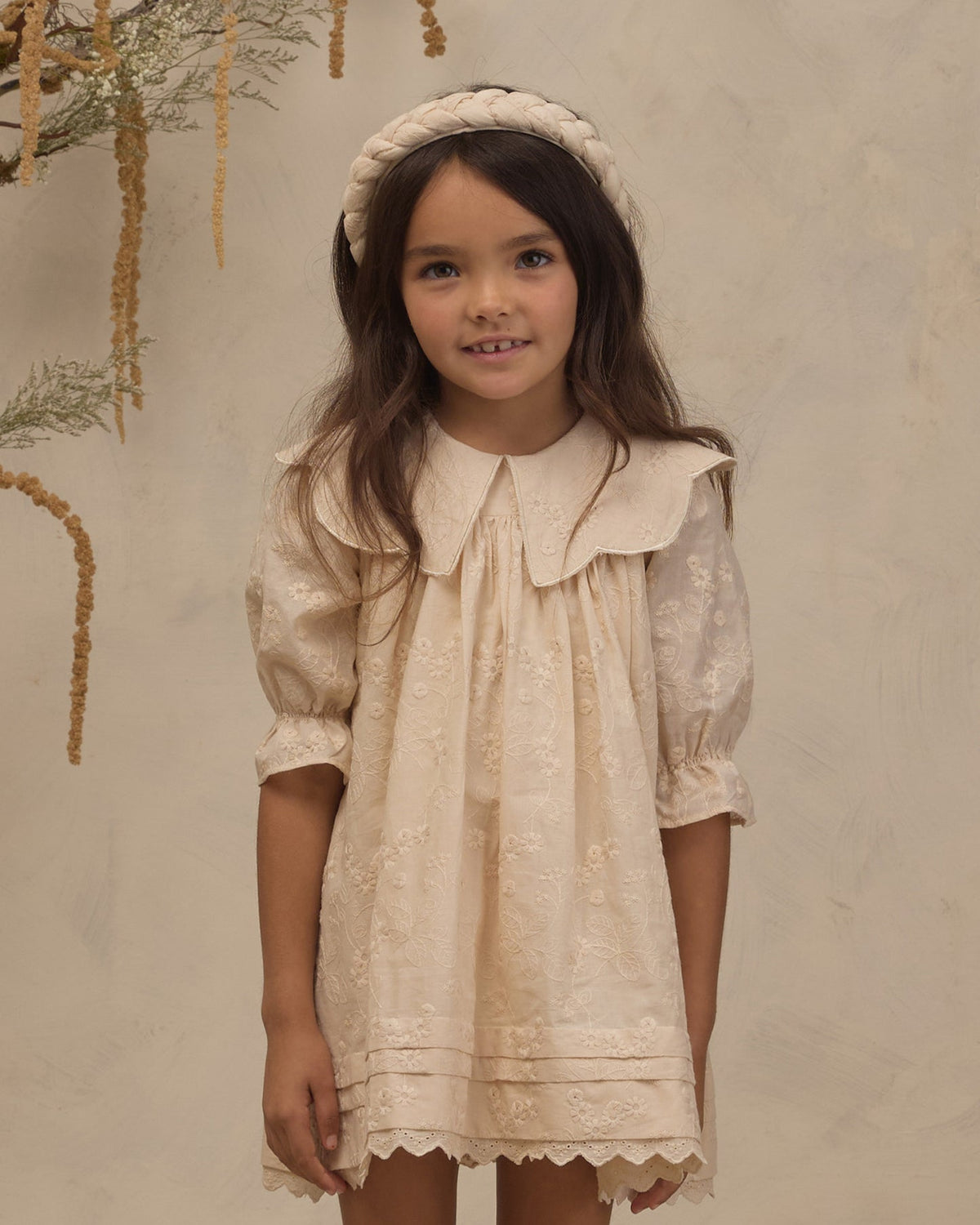 A young girl with long brown hair wearing a cream-colored lace dress with a large Peter Pan collar, puff sleeves, and a matching braided headband, standing against a textured beige wall.