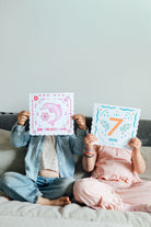 Two children sitting on a gray couch holding up colorful educational cards. The child on the left wears a denim jacket and blue jeans, holding a pink card with a dolphin illustration.