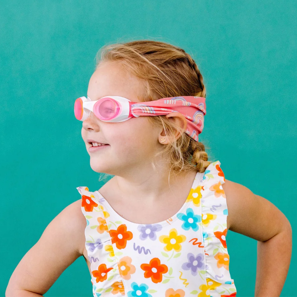 Young girl wearing colorful swim goggles and a floral swimsuit against a teal background