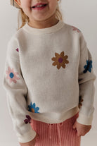 A close-up of a child wearing a cream knit sweater with colorful floral motifs in blue, pink, and brown, paired with pink knit pants against a plain light background.