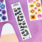 A sheet of Halloween-themed black bat stickers with white eyes and pink inner ears, displayed on a purple background alongside other sticker sheets.