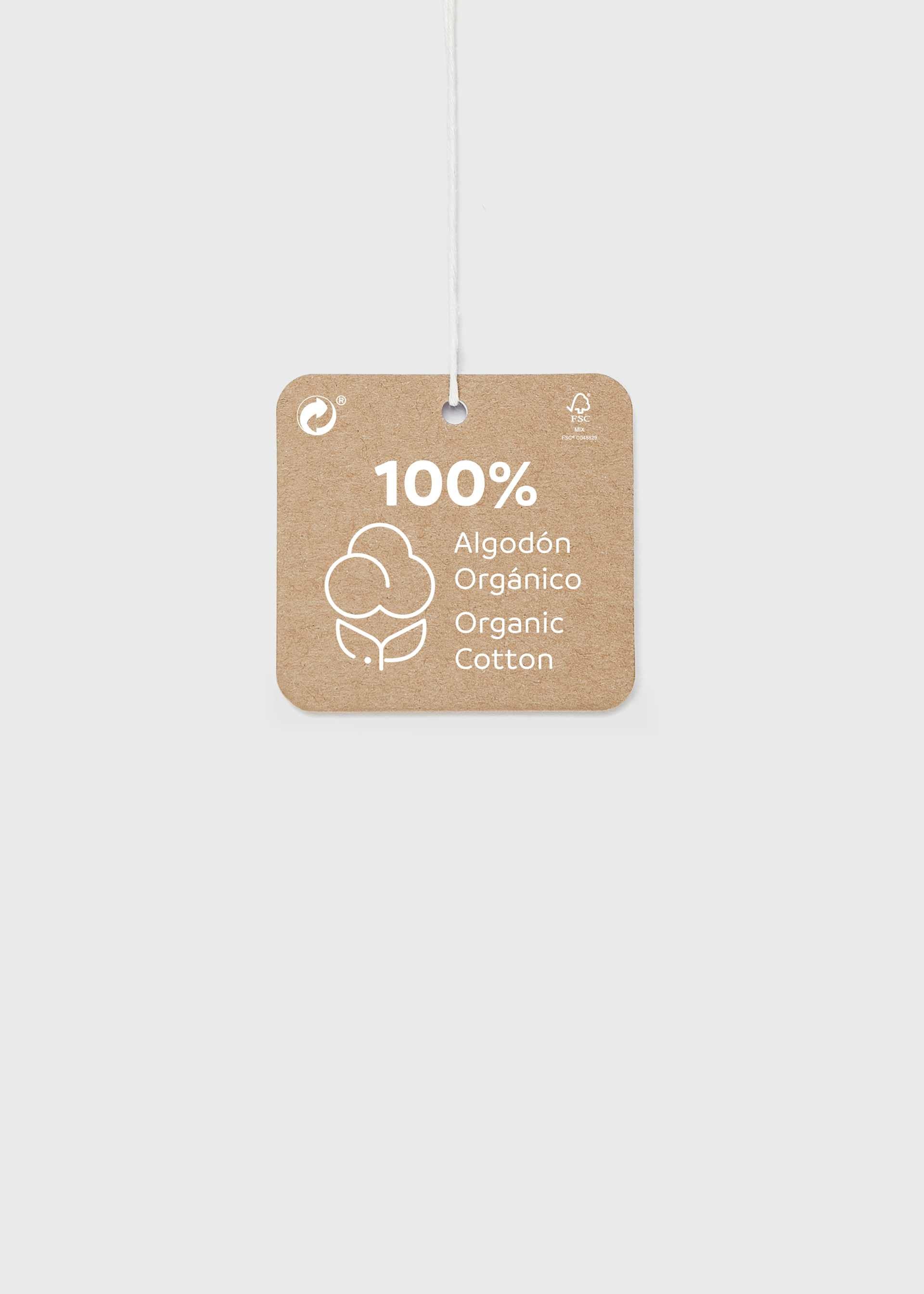A square kraft paper hanging tag with rounded corners, printed with '100% Algodón Orgánico / Organic Cotton' and a cotton boll icon, suspended by a thin white string against a light gray background.