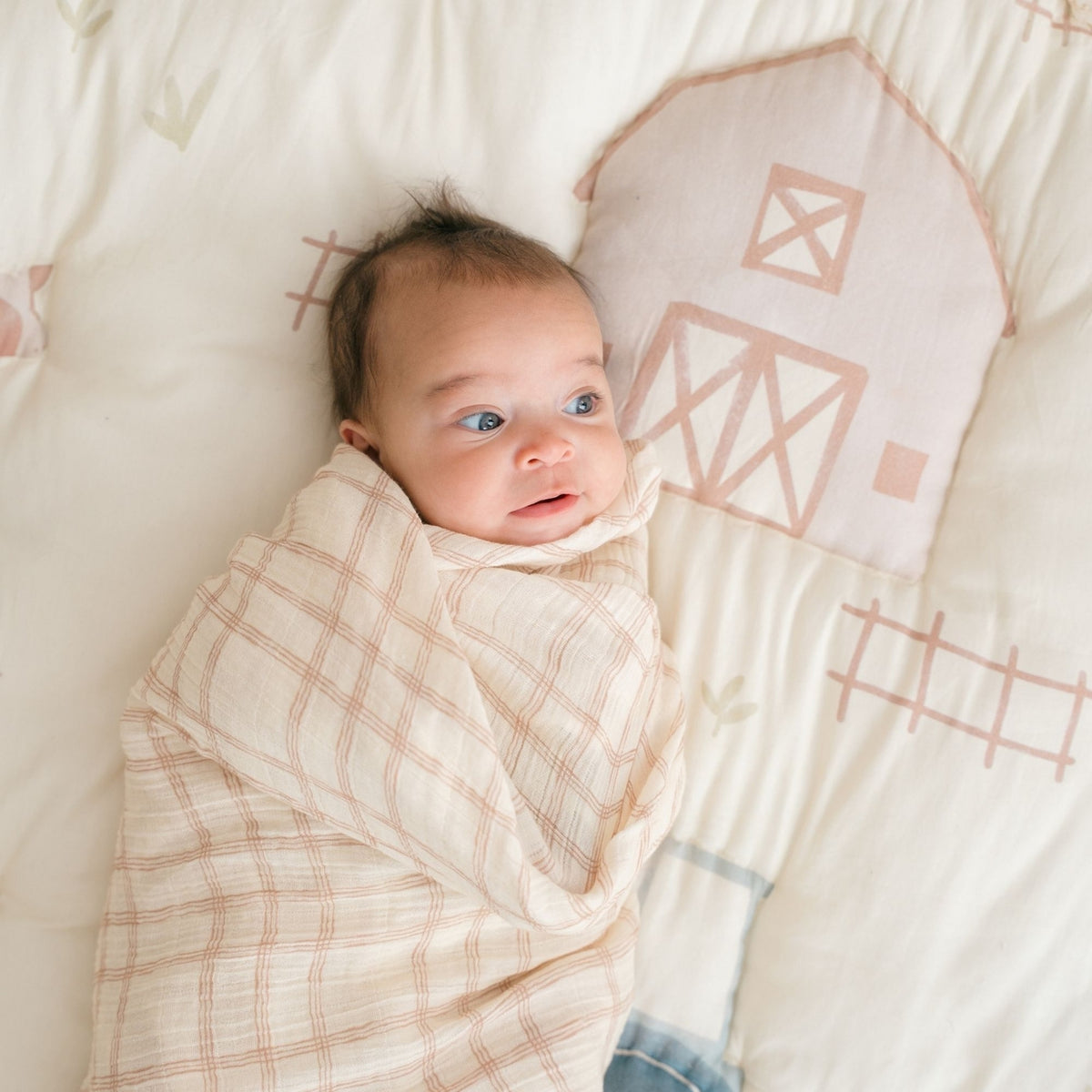 Baby wrapped in a beige swaddle blanket lying on a white crib with pink geometric patterns.
