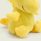 A close-up, angled view of a bright yellow Woodstock Junior by Warmies plush toy sitting upright against a plain background, focusing on its side profile, soft outstretched wing, and stitched closed eye.