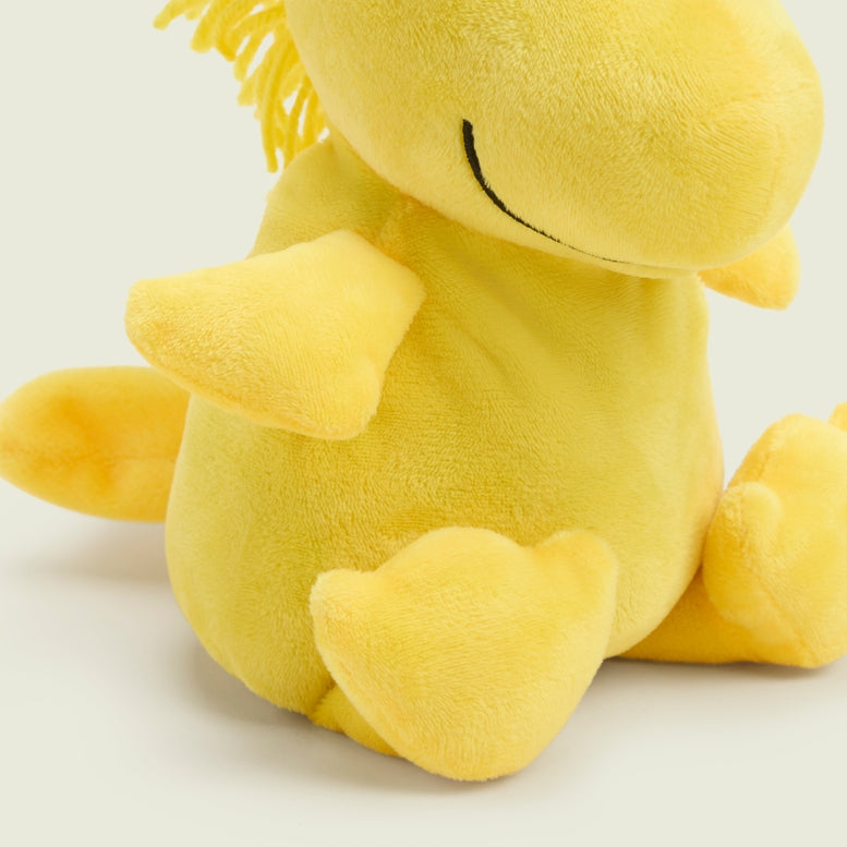 A close-up, angled view of a bright yellow Woodstock Junior by Warmies plush toy sitting upright against a plain background, focusing on its side profile, soft outstretched wing, and stitched closed eye.