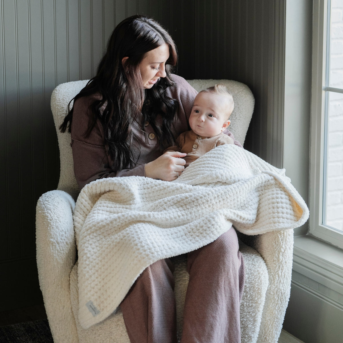 Woman holding a baby wrapped in a blanket on a chair by a window